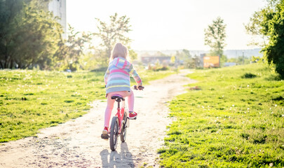 Obraz premium Cyclist preteen girl riding on bike through green sunny park, from the back