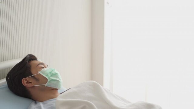 Asian patient wearing mask lying on bed in hospital facing to window.