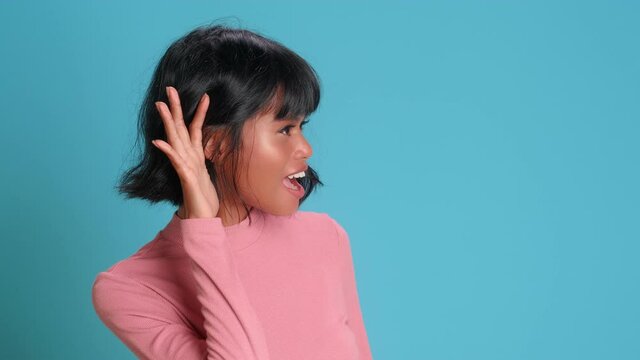 Young African Asian woman puts her hand to her ear as sign of attentive listening or eavesdropping stylish female posing on blue background. Concept of gossip and eavesdropping on important news.