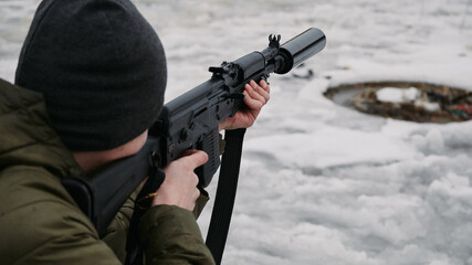 A shooter in a cap and green jacket aiming at a target with a civilian Kalashnikov-type machine gun, with a silencer in winter