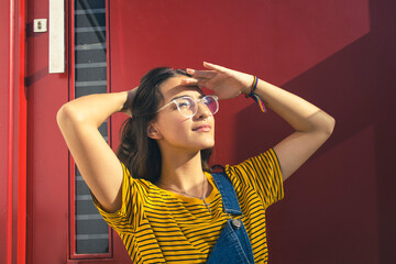 Portrait of a beautiful caucasian teenager girl wearing glasses and colorful clothes with her right hand behind her head and left hand over her eyes. Dark red door on the background. Copy space.
