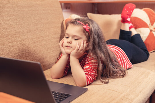 Little girl lying on sofa uses laptop for distance learning or for entertainment