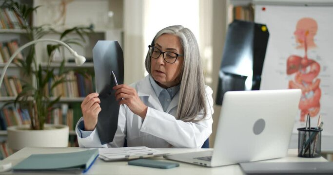 Mature woman doctor showing x ray photo and talking while having online medical consultation. Female person in glasses and white gown using laptop while communicating with patient