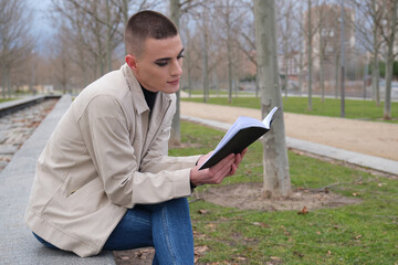 Fototapeta premium Handsome young man wearing make up, reading a book sitting on a wall. Non binary androgynous guy.