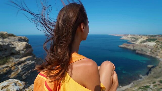 slow motion wind blowing long hair of taned traveler woman sitting on top of cliff in front of amazing seascape, bright yellow dress and hair blowing in the wind. Freedom, travel and vacation concept.