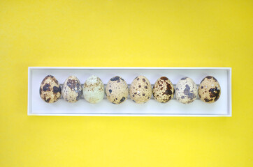 Quail eggs in a white box on a yellow background. Photo for Easter and advertising. Design, fine arts, minimalism