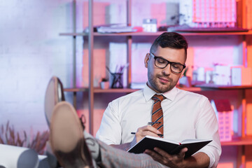 smiling architect sitting with legs on desk and writing in notebook