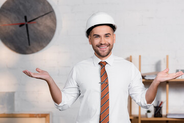 cheerful architect in helmet standing with open arms and smiling at camera