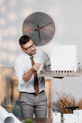 architect in eyeglasses standing at workplace and pointing with pencil at house maquette