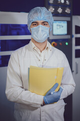 Portrait of a young man, a student, at a special electronic equipment at a factory, industrial plant, on the bottling line of a brewery in a white coat, hat, latex gloves and a folder for documents in