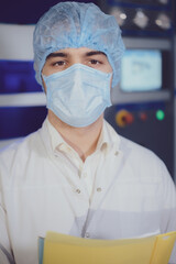Portrait of a young man, a student, at a special electronic equipment at a factory, industrial plant, on the bottling line of a brewery in a white coat, hat, latex gloves and a folder for documents in