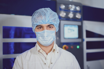 Guy wearing protective mask cap and white robe stands near control panel of modern equipment in production plant workshop closeup