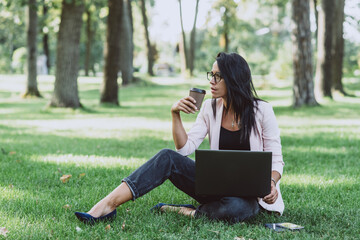 Business woman sitting in summer grass park using laptop. Works remotely during quarantine. On the street. Soft selective focus.