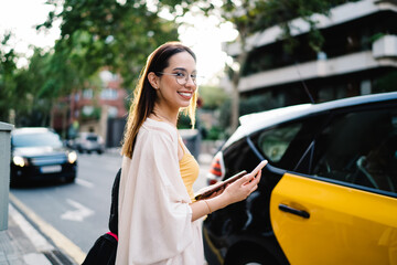Half length portrait of happy female tourist in classic spectacles using cellphone device for...