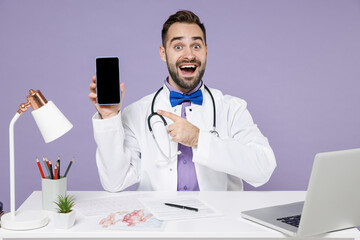 Male smiling doctor man in white medical suit sit at desk work in clinic office hold point index finger on mobile phone blank screen workspace area isolated on violet background studio portrait.