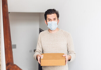 Fototapeta na wymiar A delivery guy in medical mask holding a cardboard box and looking at the camera 