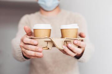 Two paper cups in a cardboard holder in man's hand. A delivery guy in a medical mask