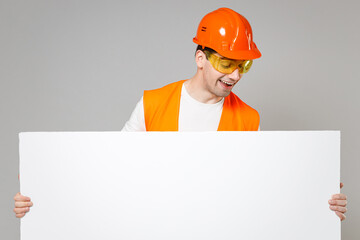 Young employee man in orange vest protective hardhat hold blank sign horizontal board with place for text isolated on grey background studio Instruments for renovation apartment Repair home concept.