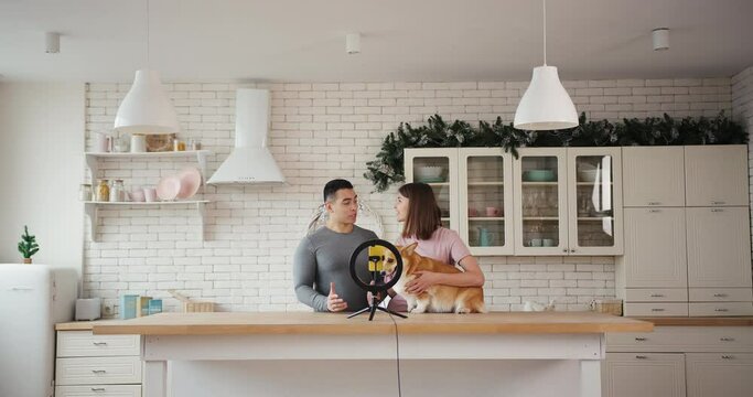 Cheerful couple with red corgi dog shoots new video for family blog with phone and light ring at table in kitchen slow motion