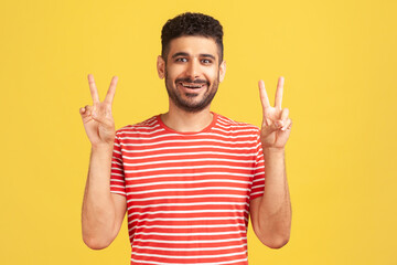 Happy successful man with beard in striped t-shirt showing v sign victory gesture with fingers, looking at camera with toothy smile. Indoor studio shot isolated on yellow background