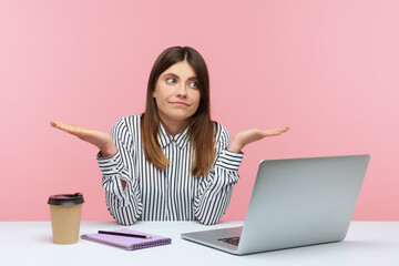 Confused uncertain woman office worker in striped shirt shrugging shoulders sitting on laptop at workplace, not sure about job completion. Indoor studio shot isolated on pink background