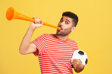 Excited bearded man in red striped t-shirt blowing in horn holding black and white soccer ball,...