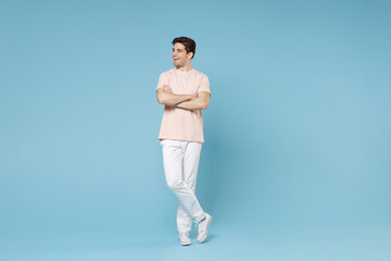 Full length of young caucasian attractive fashionable student man 20s wearing beige t-shirt white pants holding hands crossed folded looking aside isolated on blue color background studio portrait.