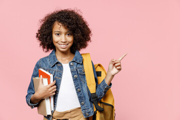 Smiling little african kid school girl 12-13 years old in casual clothes backpack hold books pointing index finger aside on copy space isolated on pastel pink background Childhood education concept