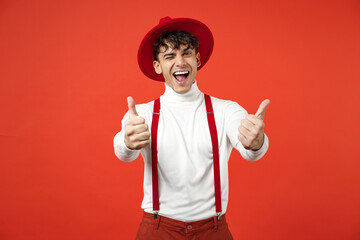 Young spanish latinos smiling happy gladden stylish fashionable student man 20s in casual hat white shirt trousers, suspenders show thumbs up gesture blink isolated on red background studio portrait.