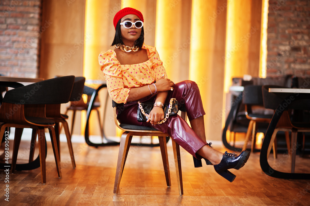 Wall mural Elegant afro american woman in red french beret, big gold neck chain, sunglasses, polka dot blouse and leather pants pose indoor.