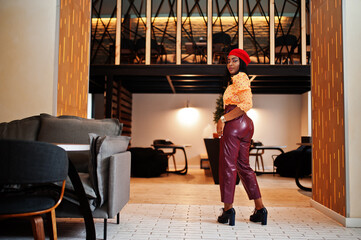 Elegant afro american woman in red french beret, big gold neck chain polka dot blouse and leather pants pose indoor.