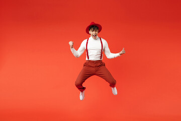 Full length of young spanish latinos overjoyed stylish man 20s in hat white shirt trousers, suspenders jumping high do winner gesture clench fist celebrating isolated on red background studio portrait