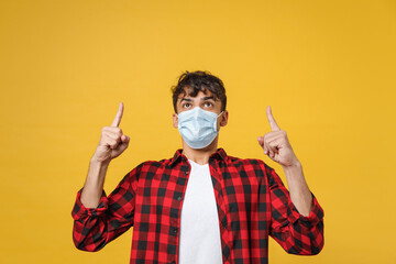 Young spanish latinos man in red checkered shirt sterile face mask coronavirus covid19 pandemic quarantine point index finger overhead on copy space isolated on yellow background studio portrait.