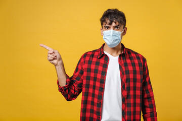 Young spanish latinos serious man in red checkered shirt sterile face mask coronavirus covid19 pandemic quarantine point index finger aside on copy space isolated on yellow background studio portrait.