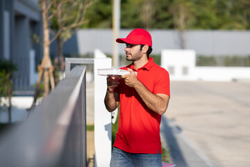 Smiling young delivery man  in red uniform holding a box give to beautiful woman costumer in front of the house.
