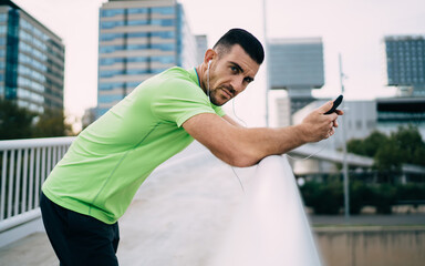 Half length portrait of male jogger in electronic headphones listening sportive audio book during morning time for cardio training, Caucasian athlete in earphones holding modern cellphone gadget