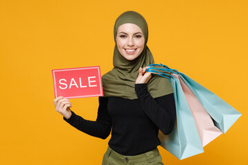 Smiling young arabian muslim woman in hijab green black clothes hold package bags with purchases after shopping sign with SALE title isolated on yellow background. People religious lifestyle concept.