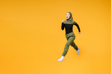 Fototapeta na wymiar Full length side view of excited young arabian muslim woman in hijab black green clothes jumping like running isolated on yellow color background studio portrait. People religious lifestyle concept.