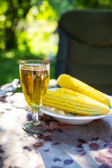 A glass of light beer in a glass along with a plate of boiled corn, standing on the table. Summer picnic, outdoor recreation.