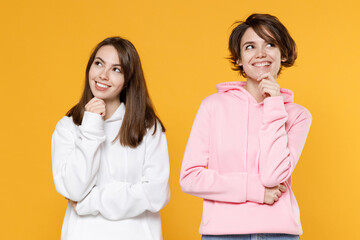 Pensive smiling funny two young women friends 20s wearing casual white pink hoodies standing put hand prop up on chin looking aside up isolated on bright yellow color wall background studio portrait.