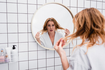 displeased woman in bathrobe adjusting hair near mirror in bathroom