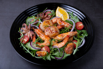 Salad with seafood, arugula, tomatoes, cucumbers, red onion and lemon on a black plate. Black background, close-up.