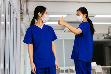 Young beautiful Asian medical nurse in scrub uniform wears face mask admire and give thumbs up to coworker at hospital during Covid19 pandemic. Trust and good cooperation in corporate culture concept