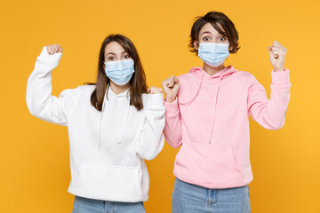 Joyful two young women friends 20s in casual hoodies sterile face mask to safe from coronavirus virus covid-19 doing winner gesture celebrating isolated on bright yellow background studio portrait.