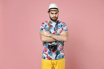 Dissatisfied young traveler tourist man in summer casual clothes hat with photo camera holding hands crossed isolated on pink background. Passenger traveling on weekends. Air flight journey concept.