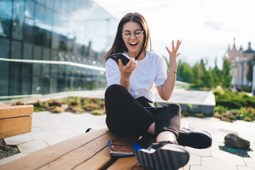 Happy female blogger in classic eyewear surprised with received amazing news using modern...