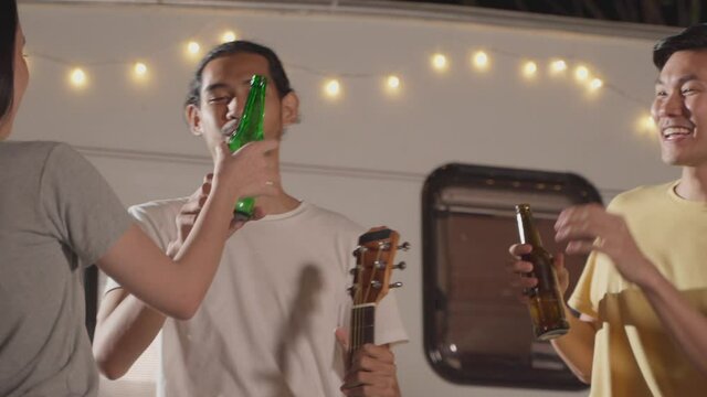 Friends enjoying outdoor guitar party at night, clink and drink beer.