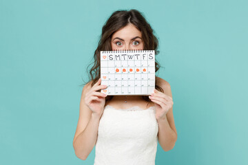 Shocked bride young woman 20s in white wedding dress cover face with female periods calendar for checking menstruation days isolated on blue turquoise background. Ceremony celebration party concept.