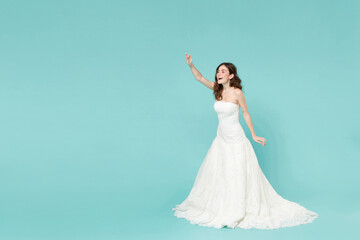 Full length of excited bride young woman 20s in white wedding dress waving and greeting with hand as notices someone isolated on blue turquoise background studio. Ceremony celebration party concept.