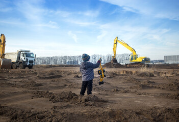 boy of 4 years old in warm clothes at a construction site with his back in the frame holds a toy...